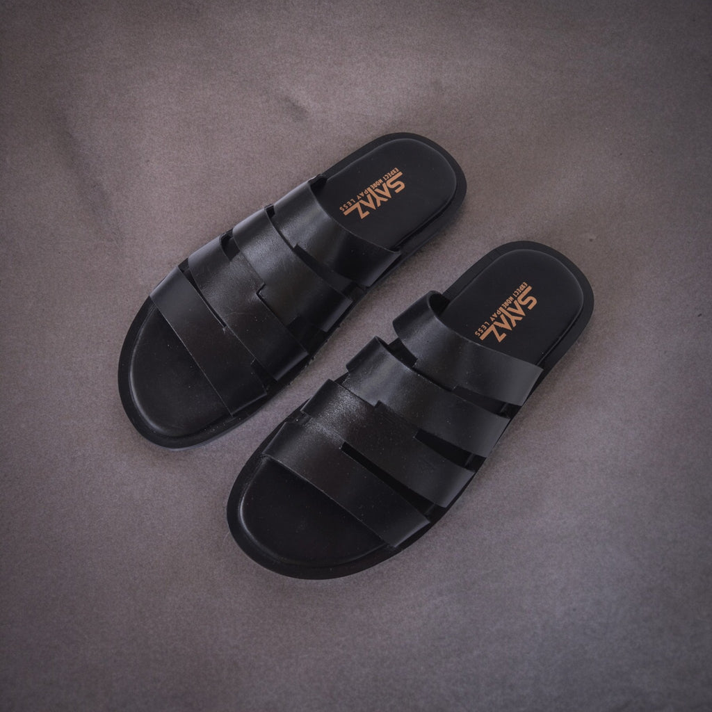 A pair of black leather slippers with a non-slip sole, placed on a textured grey surface.