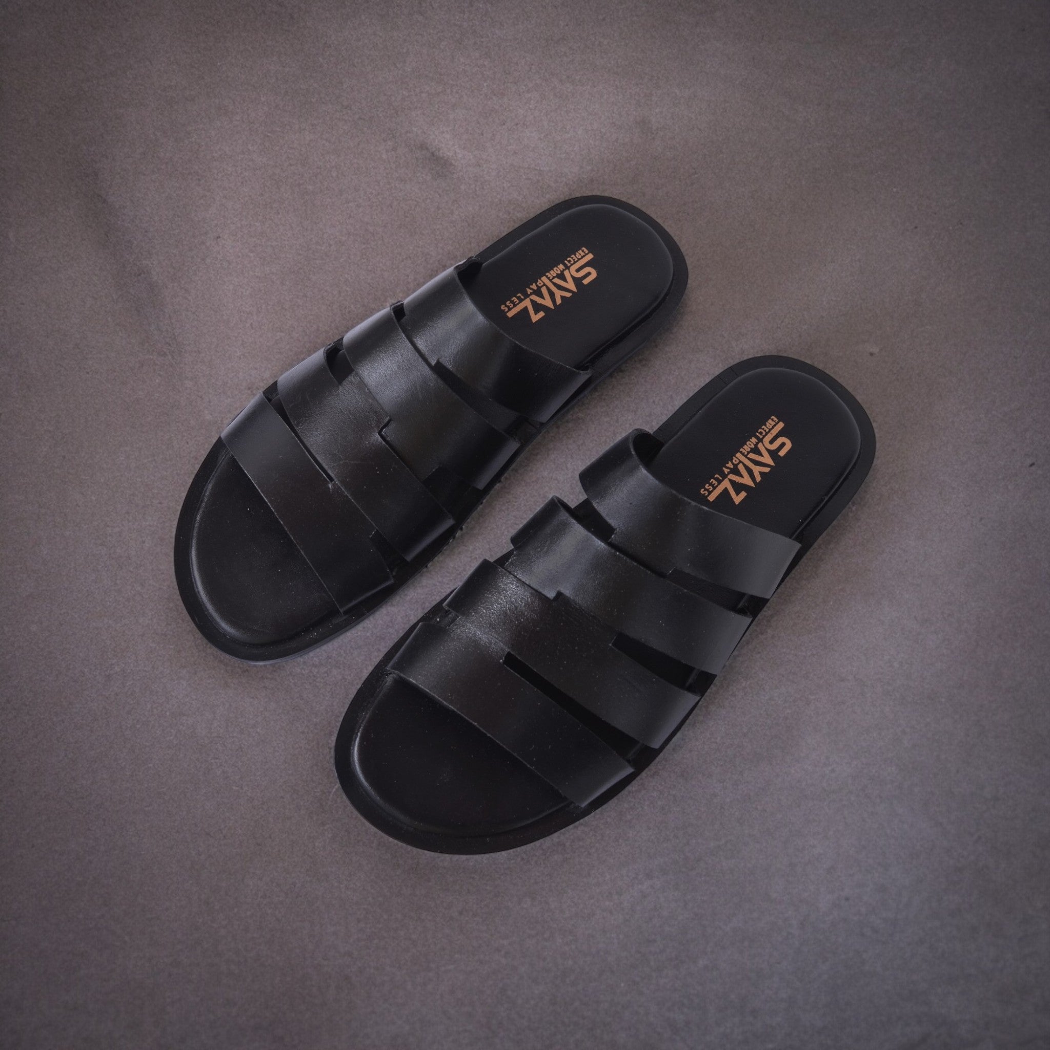 A pair of black leather slippers with a non-slip sole, placed on a textured grey surface.